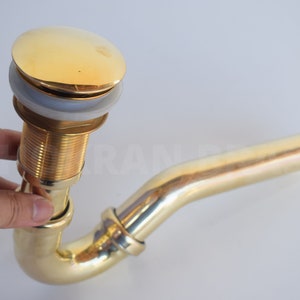 Op de afbeelding: Een gouden afvoerstop met een gebogen pijp. De stop heeft een ronde, gepolijste bovenkant en een witte rubberen afdichting.