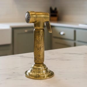 Peut inclure: Un pulvérisateur de cuisine en laiton avec une buse texturée et une poignée à levier. Le pulvérisateur a un corps cylindrique et une base évasée, posé sur un comptoir blanc.