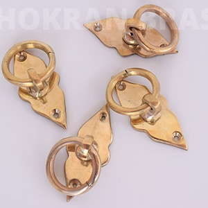 Puede incluir: Cuatro tiradores de puerta de latón con un diseño decorativo en forma de hoja. Cada tirador tiene un anillo redondo y dos pequeños tornillos.