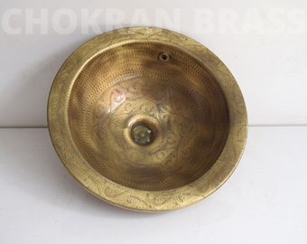Op de afbeelding: Een ronde, messing wastafel met een sierlijk, gegraveerd ontwerp. De wastafel heeft een afvoer in het midden en een klein gaatje aan de rand.