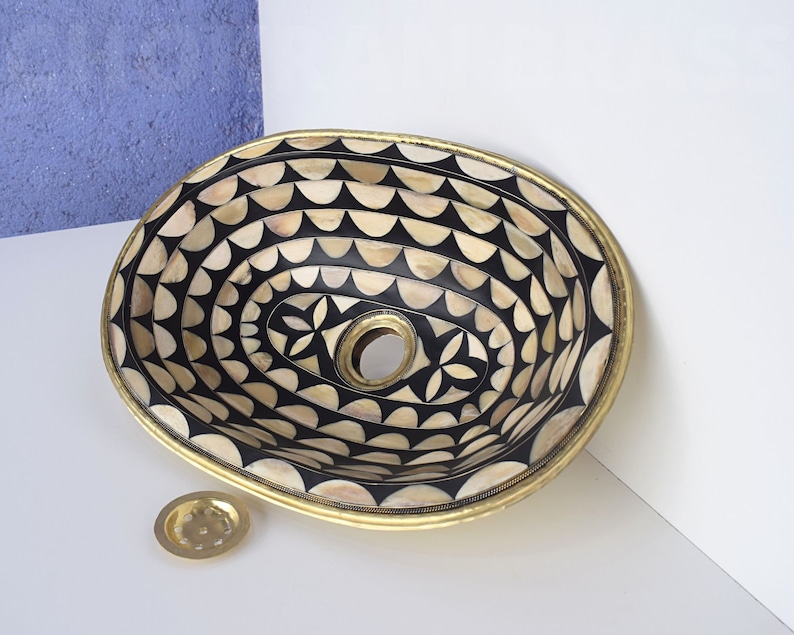 Op de afbeelding: Ovaalvormige wastafel gemaakt van zwart en wit botinleg met een gouden rand. De wastafel heeft een geometrisch patroon en een afvoergat in het midden.