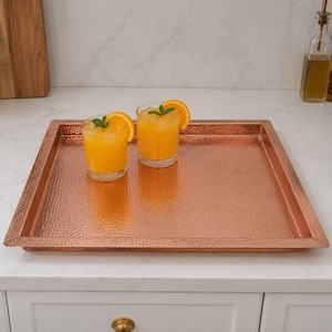 Peut inclure: Un plateau carré en cuivre martelé avec une finition métallique. Deux verres transparents remplis de jus d'orange, de glaçons et de tranches d'orange avec de petites feuilles de menthe verte sont posés sur la surface du plateau en cuivre.