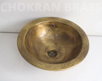 Op de afbeelding: Een ronde, gouden messing gootsteen met een sierlijk ontwerp rond de rand. De gootsteen heeft een klein afvoergat in het midden.