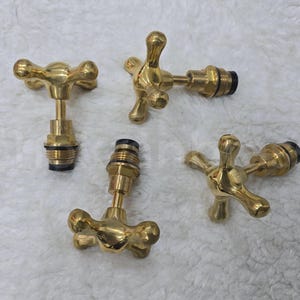 May include: Four brass faucet handles with a cross-shaped design. The handles are gold-colored and appear to be vintage or antique. The handles are on a white, textured surface.