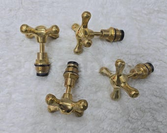 May include: Four brass faucet handles with a cross-shaped design. The handles are gold-colored and appear to be vintage or antique. The handles are on a white, textured surface.