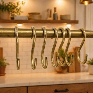 May include: A set of seven brass-colored S-hooks hanging from a brass rod. The hooks are curved and designed for kitchen or home organization. The background shows a kitchen setting with wooden shelves and a white tiled backsplash.