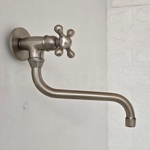 Pode incluir: Uma torneira de parede prateada com uma pega em forma de cruz. A torneira tem um bico curvo e uma base redonda.