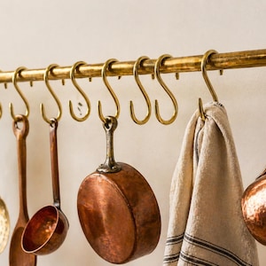 Può includere: Una barra da cucina color ottone con ganci a S che sostengono vari utensili da cucina in rame e legno, tra cui un mestolo, un cucchiaio e una piccola padella. Un canovaccio di lino a righe è appeso a uno dei ganci. Il tutto è su sfondo neutro.