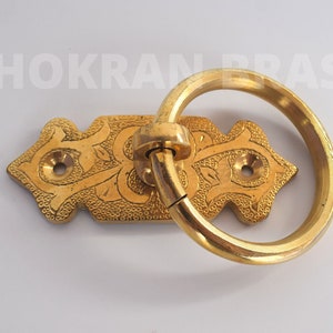 May include: Gold-toned metal ring pull handle with an ornate design. The handle is attached to a rectangular plate with a decorative pattern.