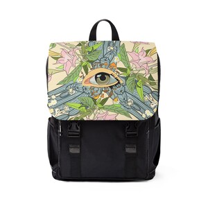 May include: Black backpack with a flap featuring a colorful design of an eye, flowers, and mushrooms. The eye is centered within a floral and geometric pattern. The backpack has adjustable straps and a top handle.