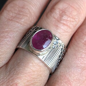 May include: A silver ring with a large, oval-shaped ruby gemstone. The ring has a textured band with vertical lines and a dotted pattern on the sides. The ruby is a deep red color.