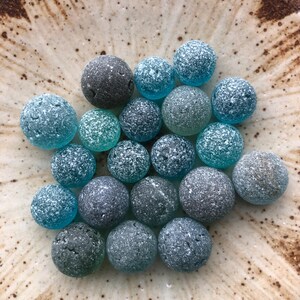 以下が含まれることがあります： 青、ティール、グレーの色合いの、小さくて丸い装飾的なガラスビーズのコレクション。 ビーズはテクスチャ加工されたつや消し外観で、まだらのセラミックプレートに配置されています。 ビーズのサイズと色は異なります。