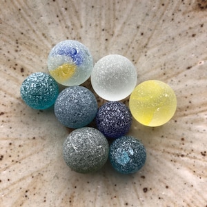 以下が含まれることがあります： 青、緑、黄色の様々な色合いの、8つのカラフルなガラス玉のコレクション。玉はまだら模様のテクスチャで、まだら模様の薄茶色のボウルに配置されています。玉は様々なサイズです。