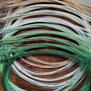 May include: A collection of thin, metallic-finish cords arranged in concentric circular loops. The cords feature a gradient of colors including gold, silver, light green, and dark green, with a shimmering texture visible along each individual strand.