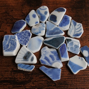 Puede incluir: Un conjunto de fragmentos de cerámica azul y blanca con diversos diseños, incluyendo motivos florales y de olas, dispuestos sobre una superficie de madera oscura. Los fragmentos, de diferentes tamaños y formas, crean una composición similar a un mosaico.