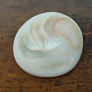 May include: A smooth, circular, white cabochon with a polished finish. The surface features swirling, translucent patterns in soft shades of pale green, cream, and light peach, creating a subtle, iridescent effect across the rounded, dome-like shape.