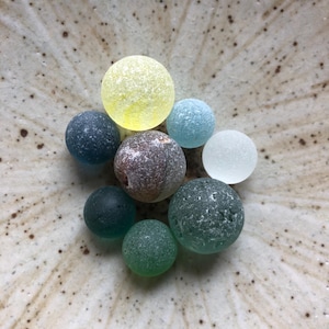 以下が含まれることがあります： 黄色、青、緑、グレーの色合いの、カラフルな丸いガラス玉のコレクション。 ガラス玉は、斑点のある薄茶色の模様の陶器のボウルに配置されています。