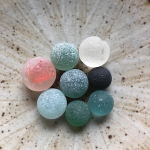 以下が含まれることがあります： ピンク、グリーン、ブルー、ブラックの色合いの、カラフルな丸いガラス玉のコレクション。ガラス玉は、まだらの薄茶色の陶器のボウルの中に装飾的なパターンで配置されています。ガラス玉は、つや消しで質感のある外観をしています。