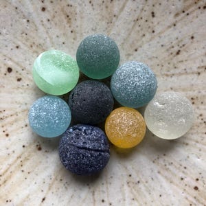 以下が含まれることがあります： 緑、青、黄色、グレーの色合いの、9つのカラフルな丸いガラスビーズのコレクション。ビーズはフロスト加工された質感で、円形に配置されています。まだらのライトブラウンのセラミック表面に表示されています。