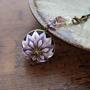 Puede incluir: Un colgante redondo hecho a mano con un diseño floral detallado en tonos morado, blanco y dorado. El colgante está sujeto a un cordón negro con cuentas decorativas y un cierre. El fondo es una superficie de madera.