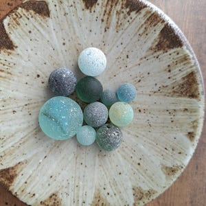 以下が含まれることがあります： 青、緑、グレーの色合いのカラフルなガラス玉のコレクションが、装飾的な陶器の皿に並べられています。 皿はクリーム色のベースに茶色の花の模様が縁に施されています。 ガラス玉はサイズと質感が異なります。