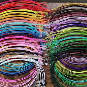 Puede incluir: Una colección de alambres para manualidades finos y flexibles dispuestos en bobinas circulares. El surtido presenta un amplio espectro de colores que incluye rojo, amarillo, naranja, turquesa, rosa, blanco, marrón, morado, verde lima y negro.