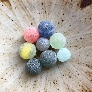 以下が含まれることがあります： ピンク、青、緑、黄色、グレーの色合いの、カラフルで質感のあるガラス玉のコレクション。 斑点のある薄茶色の陶器のボウルの中に、円形に配置されています。