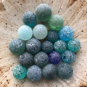 以下が含まれることがあります： さまざまな青、緑、グレーの色合いのシーグラスマーブルのコレクションが、まだらのセラミックプレートに配置されています。マーブルはフロスト加工された質感で、まだらの模様がいくつかあります。プレートは茶色とベージュの色合いです。