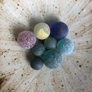以下が含まれることがあります： 紫、黄色、青、緑の色合いの、カラフルで質感のあるガラス玉のコレクション。 ガラス玉は、斑点のあるクリーム色のセラミックボウルの中に円形に配置されています。 ガラス玉はサイズと質感が異なります。