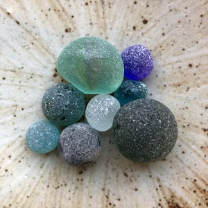 以下が含まれることがあります： さまざまな色合いの、滑らかで丸みを帯びたシーグラスのコレクション。ティール、ブルー、グレー、クリアなどがあり、まだらのタン色のボウルに配置されています。シーグラスはすりガラスのような外観をしています。