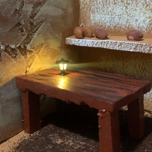 May include: A small, black, metal lantern with a light inside sits on a wooden table with a red-brown finish. The table has a rough, rustic look and is made of wood planks. The lantern is casting a warm glow on the table.