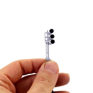 Può includere: Un modello in miniatura di un segnale ferroviario con tre luci nere. Il segnale è grigio e ha una piccola scala laterale. Il modello è tenuto in mano da una persona su sfondo bianco.