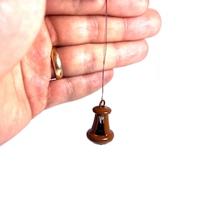 Può includere: Una piccola lampada a sospensione in ceramica marrone con un filo rosso. La lampada è appesa a una mano.