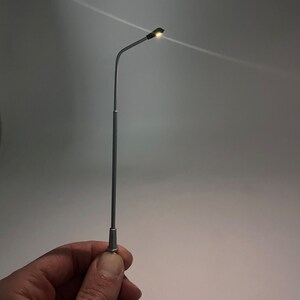 K&ouml;nnte beinhalten: Eine silberne Modell-Stra&szlig;enlaterne mit einem schwarzen Leuchtenk&ouml;rper und einem kleinen, warmwei&szlig;en LED-Licht.