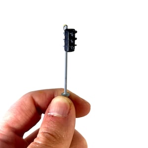Può includere: Un semaforo nero in miniatura con tre luci di segnalazione, montato su un palo grigio. La base del palo è un piccolo disco rotondo color argento. Il semaforo è tenuto in mano da una persona su sfondo bianco.
