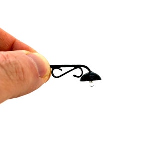 Può includere: Miniatura di lampione da strada in metallo nero con un design decorativo a volute e un paralume nero a forma di cupola. La lampada ha una piccola lampadina trasparente.