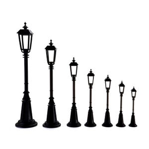 May include: A collection of seven black miniature street lamps of varying heights. Each lamp features a classic design with a square lantern top, a decorative base, and a slender pole. The lamps are arranged in a row against a white background.