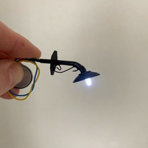 Può includere: Un piccolo lampione da strada modello nero con una luce LED bianca. Il lampione è fissato a un supporto a parete e ha un braccio curvo. Il lampione è cablato a un pacco batteria.