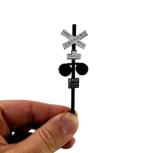 Puede incluir: Señal de cruce de ferrocarril en blanco y negro con el texto "RAIL CROSSING ROAD" y "2 TRACKS". La señal está montada en un poste negro con dos luces negras a cada lado de la señal. Un letrero blanco debajo de las luces dice "STOP ON RED SIGNAL".