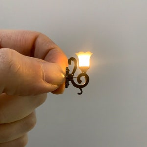 Puede incluir: Una lámpara de pared de metal negro en miniatura con una bombilla de vidrio blanco que está encendida. La lámpara tiene un diseño decorativo de desplazamiento.