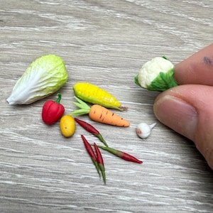 May include: Miniature vegetables including a head of lettuce, red bell pepper, yellow corn, carrot, red chili peppers, a lemon, a head of cauliflower, and a garlic clove. The vegetables are arranged on a light wood-grain surface.