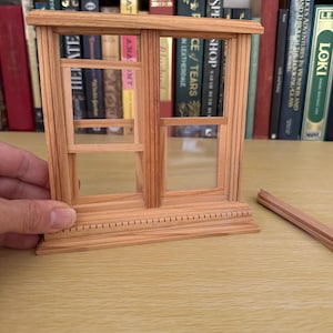 Op de afbeelding: Een miniatuur houten raamkozijn met twee ruiten en een decoratieve basis. Het frame is gemaakt van lichtgekleurd hout en is gedeeltelijk gemonteerd. Een klein stukje hout ligt ernaast. Het raam staat tegen een achtergrond van boeken.