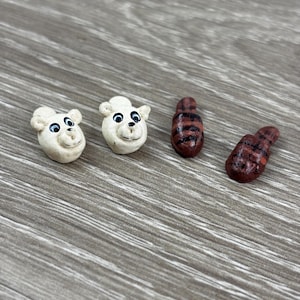 May include: Four small, handmade polymer clay figurines. Two are white bear heads with black eyes and smiles. The other two are brown and red striped, worm-like shapes. The figurines are arranged on a gray wooden surface.