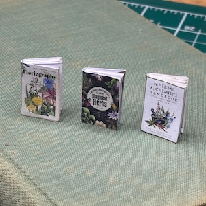 以下が含まれることがあります： 花と植物のイラストが描かれたミニチュアの書籍が3冊。書籍のタイトルは「Floriography」、「Magickal Herbs」、「The Herbal Alchemist's Handbook」です。