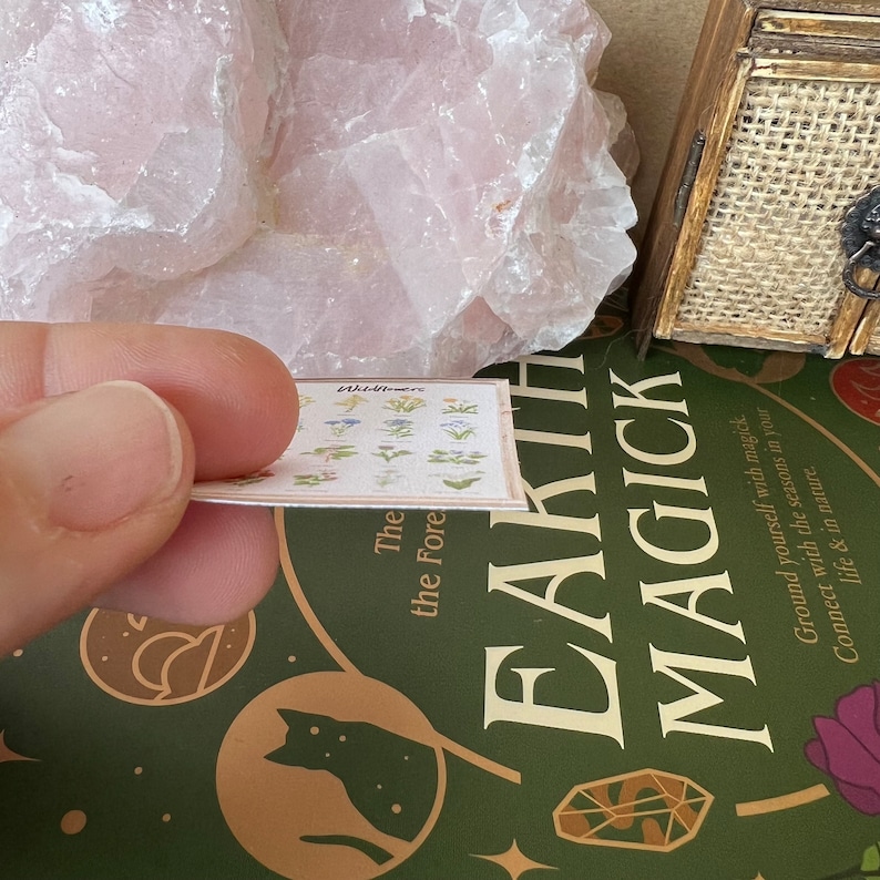 May include: A close-up of a small card with a drawing of wildflowers and the text "Wildflowers" and "The Forest". The card is being held by a hand and is resting on a green book with the text "Earth Magick" on the cover.