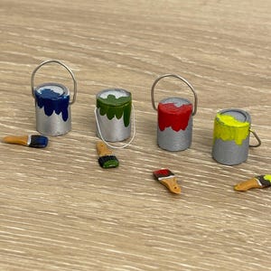 Puede incluir: Miniaturas de botes de pintura plateados con pintura azul, verde, roja y amarilla. También hay pequeños pinceles con colores a juego. Los artículos están dispuestos sobre una superficie de madera clara, lo que sugiere un tema de artesanía o arte.
