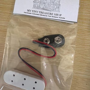 May include: A clear plastic bag containing a black battery connector with red and black wires, and a white connector with five holes. The bag has a white label with the text "MY TINY TREASURE SHOP" and a drawing of a house.