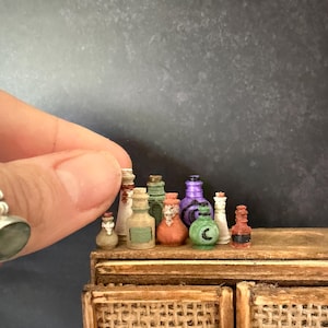 May include: A collection of ten miniature bottles in various shapes and colours, including green, purple, orange, white, and brown, arranged on a wooden surface. Some bottles feature small painted details like a black crescent moon or labels.