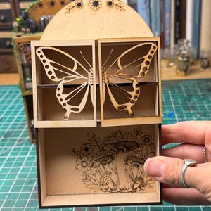 May include: A wooden display shelf featuring butterfly cutouts and a mushroom and floral design. The shelf is finished in light brown and is designed for small items. The butterfly cutouts are finely detailed.