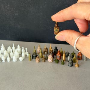 May include: A collection of miniature apothecary bottles in various colours including white, brown, green, and black. The bottles feature diverse shapes, some with stoppers and labels, arranged in a group with one brown bottle held up.
